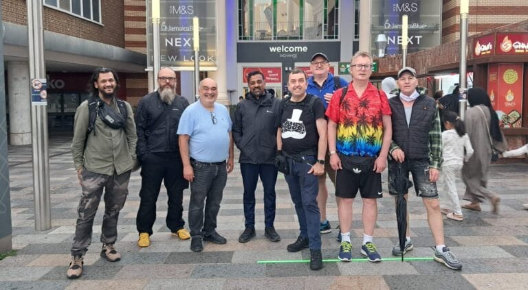 A group of men dressed in sports attire looking at the the camera and smiling outside the Ilford Station entrance of the Exchange Shopping Centre.