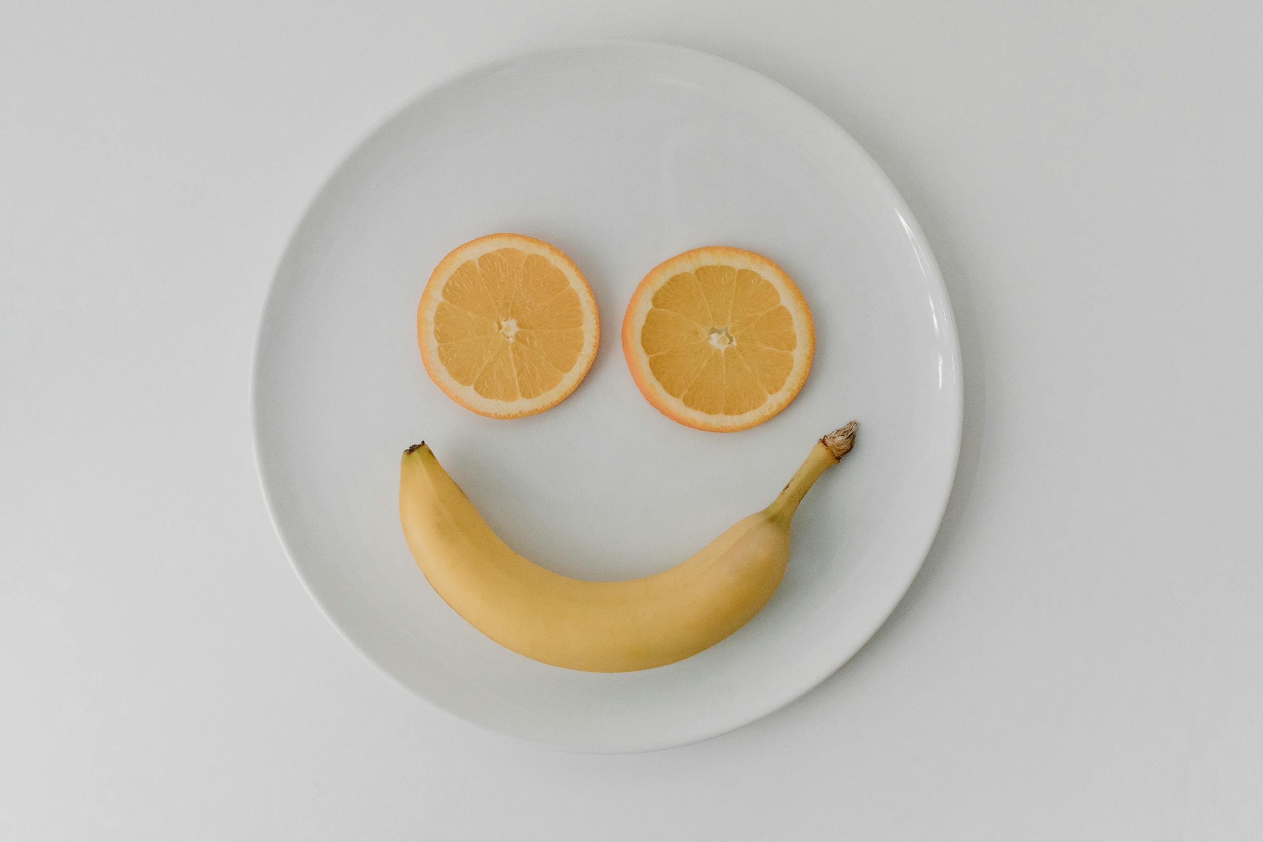 A playful smiley face made with a banana and orange slices on a ceramic plate, viewed from above.